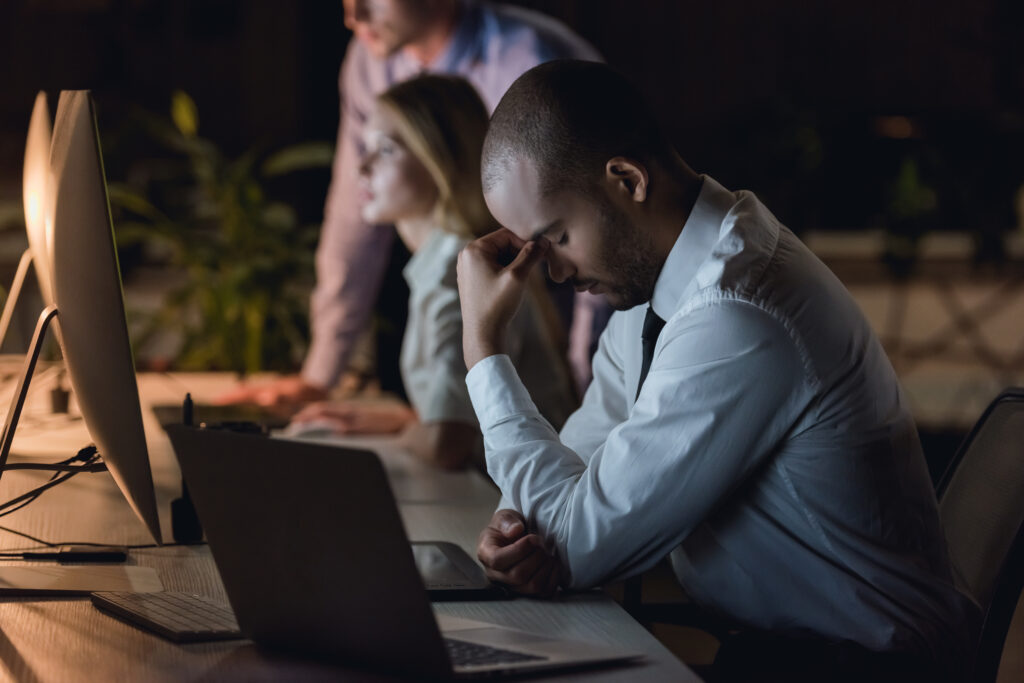 Tired Afro American businessman is massaging his nose bridge while working with a computer in office late in the evening, his colleagues near.
