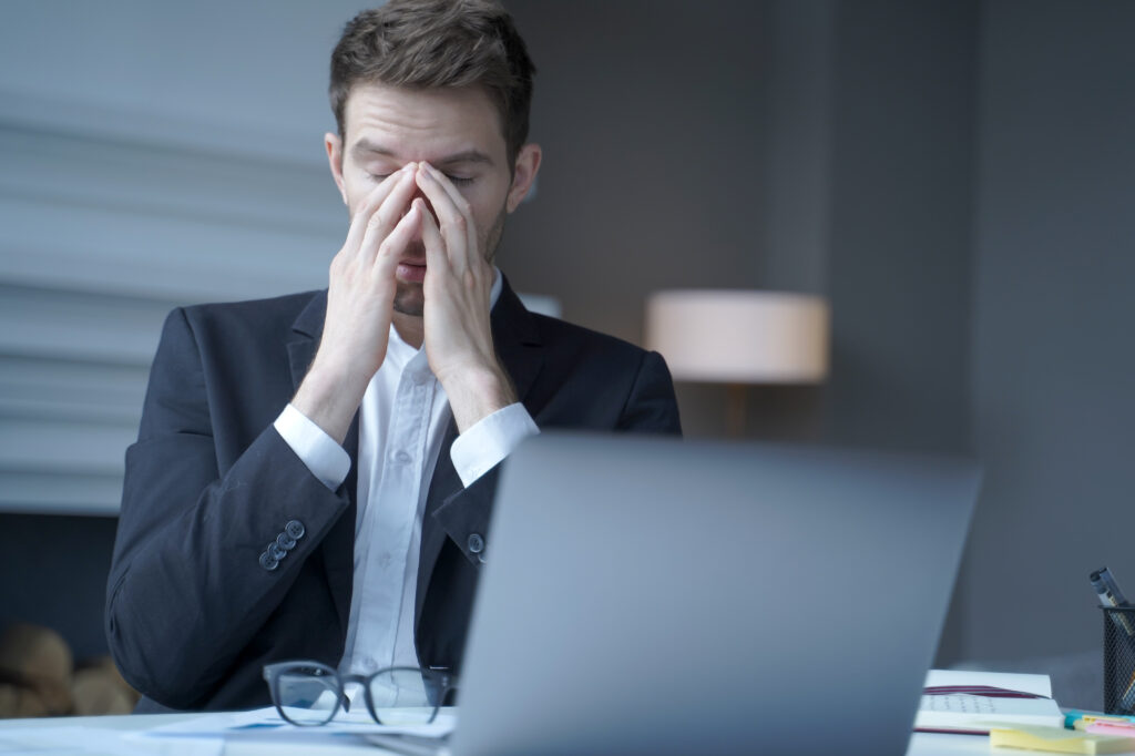 Overworked male entrepreneur sits at home office while took off glasses massages bridge of his nose with eyes closed, exhausted man suffering from heavy headache and fatigue. Health problem at work.